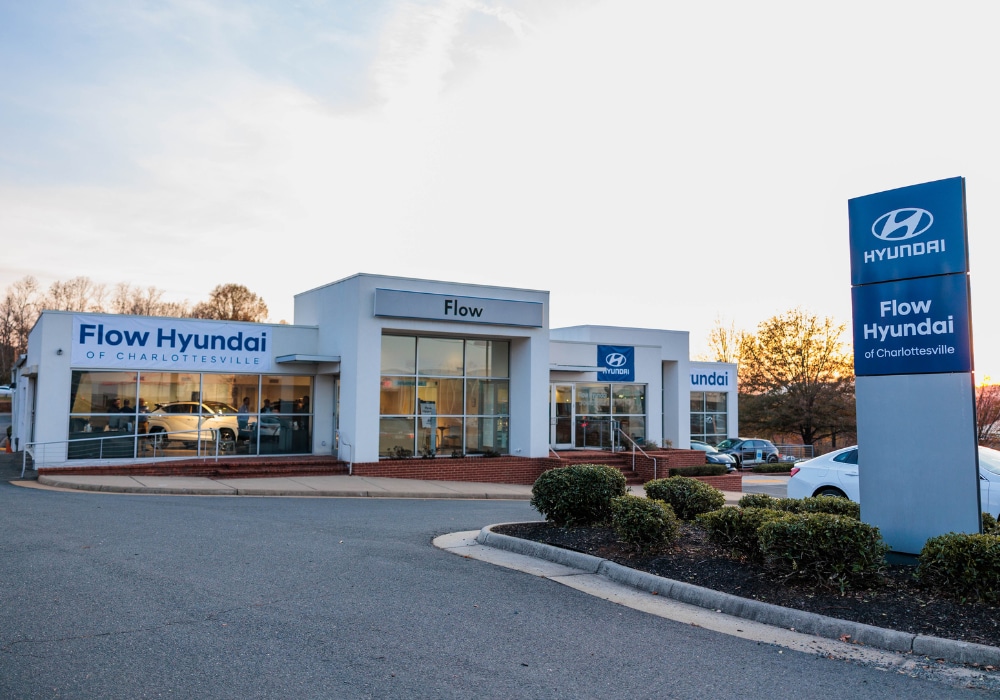 Exterior shot of the Flow Hyundai of Charlottesville car dealership. There are cars parked outside the building and a sunset in the background