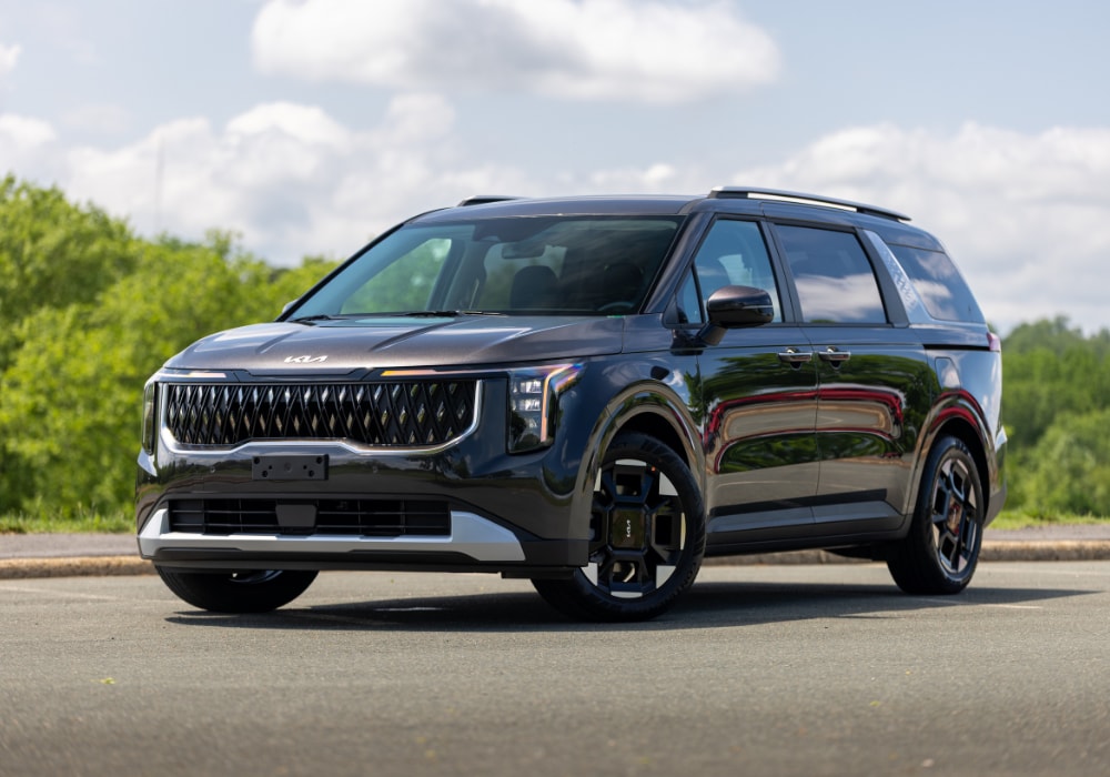 2025 Kia Carnival for sale parked on a paved lot. The car is black and there are trees in the background