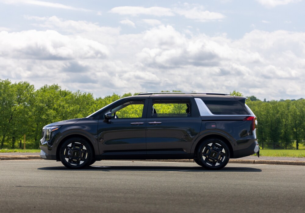 Exterior view of the driver's side of a Kia Carnival parked in a paved lot with trees and clouds in the background