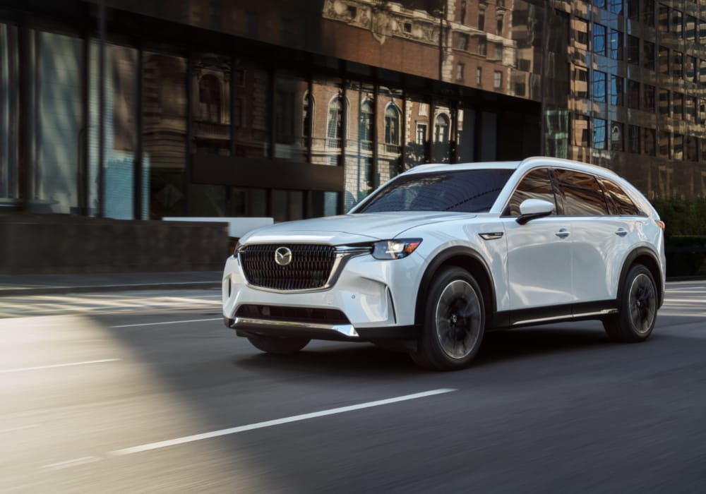 new-mazda-cx-90-phev.png White 2025 Mazda CX-90 Hybrid driving down a street with a large building in the background