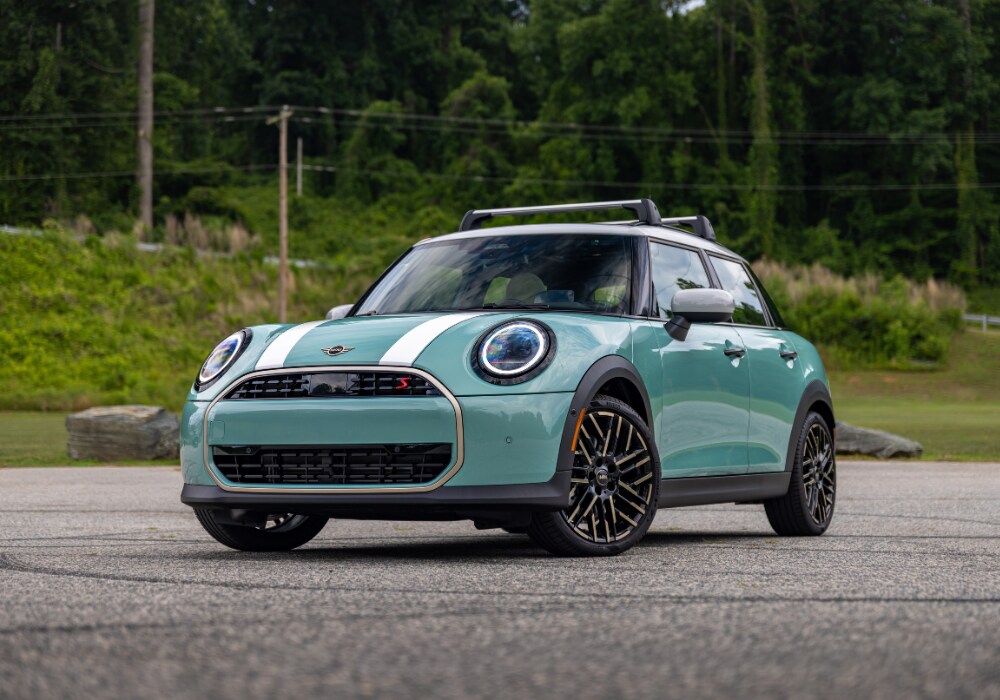 Front view of a new Mini Cooper 4 door parked in front of trees. The car is teal with white and gold details