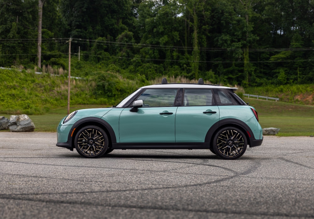 Passenger side view of a 2026 4 Door Mini Cooper. The car is teal and there are trees in the background