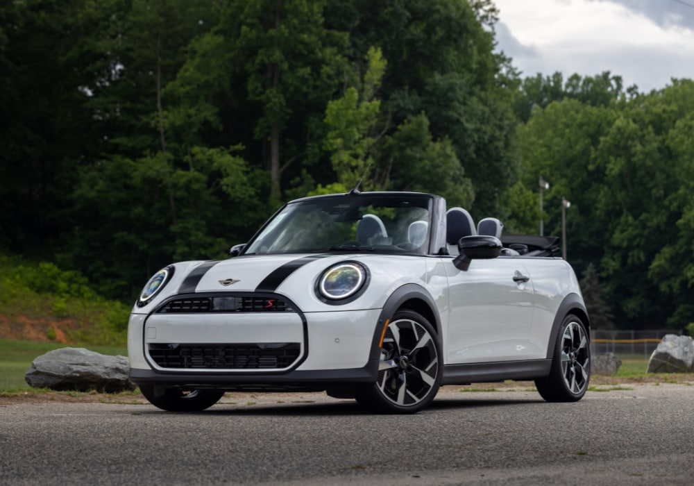 Front view of a 2026 Mini Cooper Convertible. The car is white with black stripes and there are trees in the background
