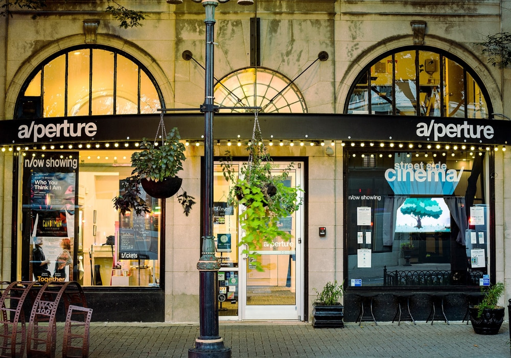 Street view of the a/perture movie theater. The building is lit up and there are movie posters hanging from the windows