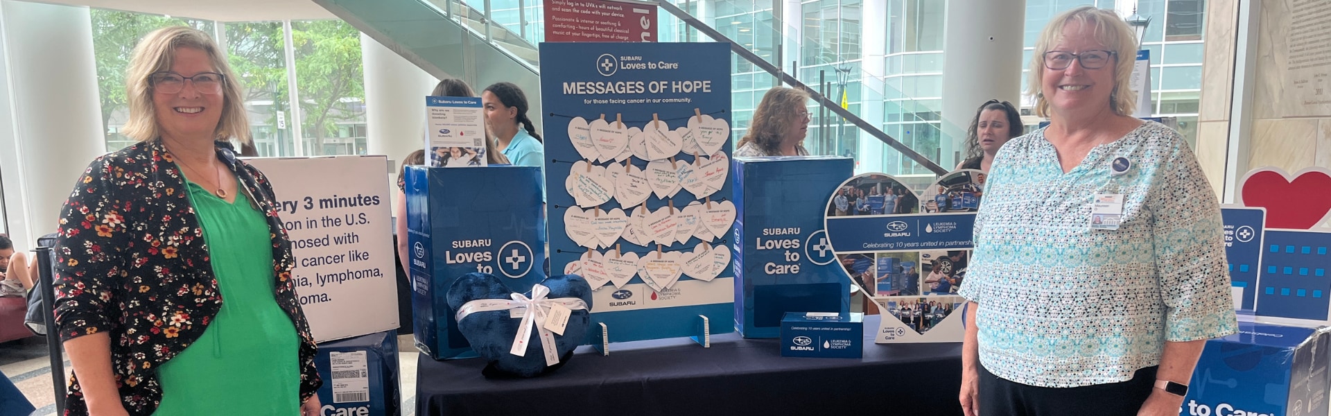 Two woman stand next to a Subaru Loves to Care table display in a hospital lobby