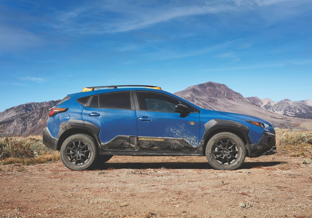 Passenger side view of a Subaru Crosstrek. The SUV is blue and there are mountains in the background