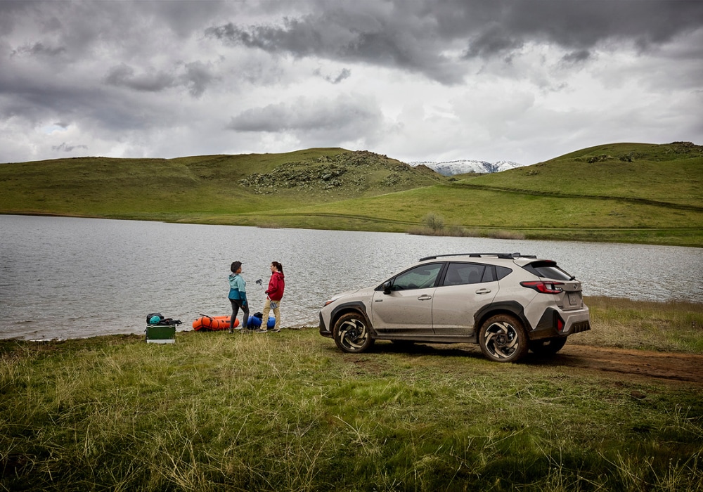 2026 Subaru Crosstrek Hybrid parked besides a large body of water with mountains in the background. There are two people standing next to the car