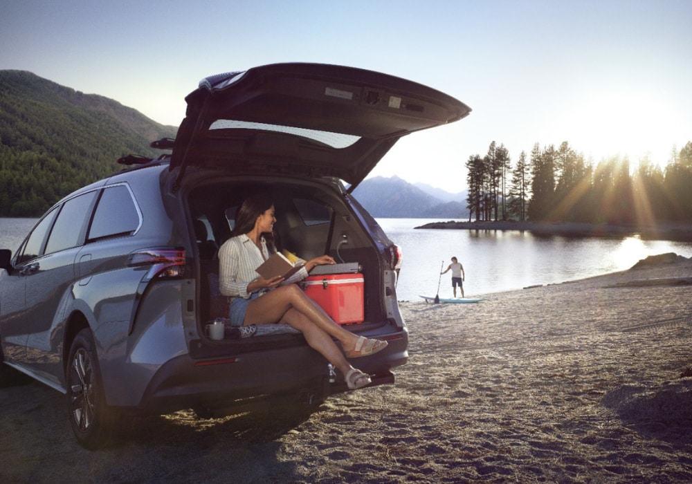 toyota-sienna-for-sale.png A woman sitting in the open truck of a Toyota Sienna. The minivan is parked in front of a lake