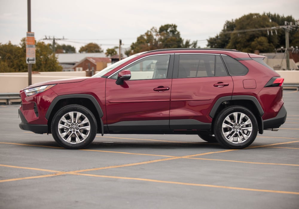Driver's side exterior view of a new Toyota RAV4 for sale, parked in a parking lot with trees and buildings in the background