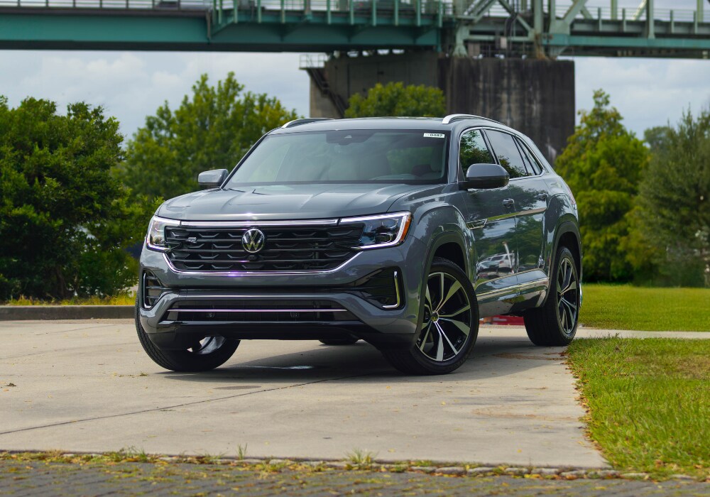 Dark blue 2025 Volkswagen Atlas Cross Sport SUV parked on a paved surface with a bridge in the background