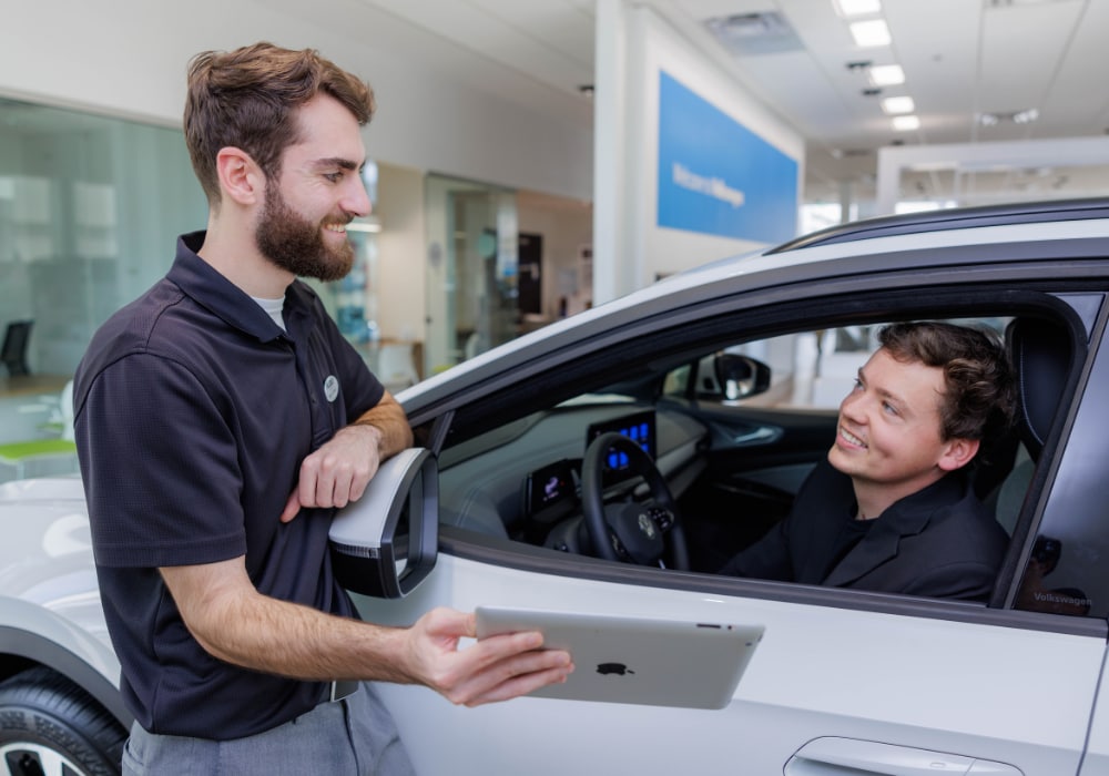A Flow employee assisting a customer who is sitting inside a car at the Flow Volkswagen of Asheville dealership. The employee is smiling and showing the customer a tablet screen