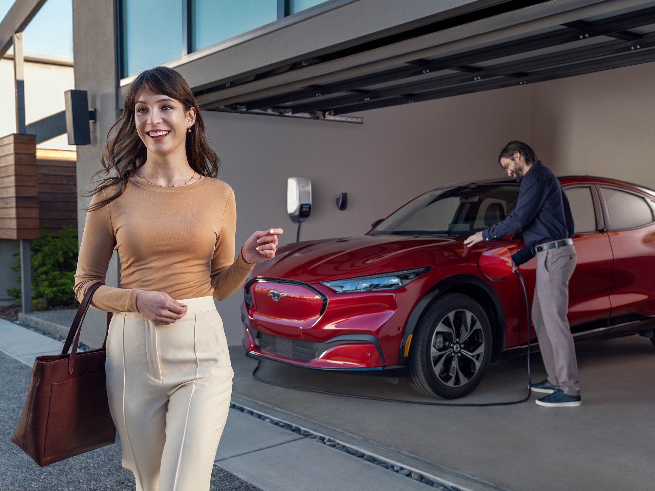A woman walks away from a Mustang Mach-E as a man plugs it into a home charging station