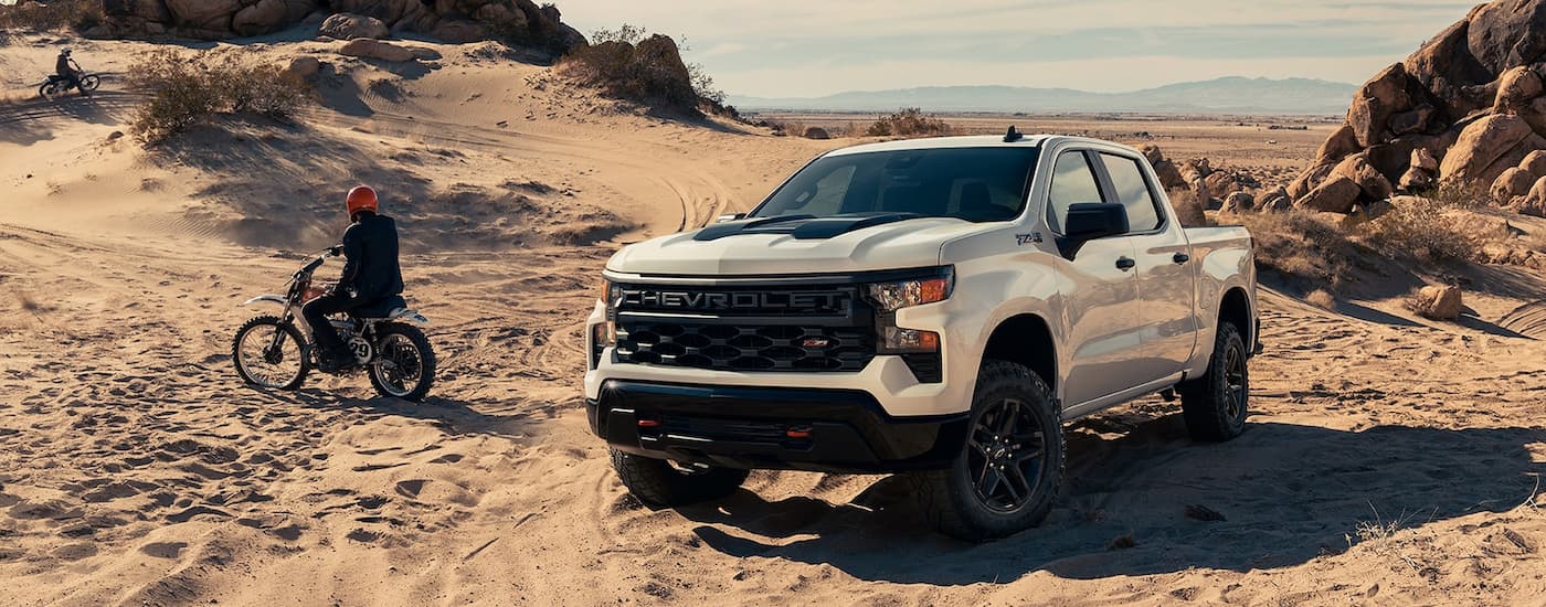People off-roading near a white 2022 Chevy Silverado 1500 Z71 Trailboss parked in sandy dunes.