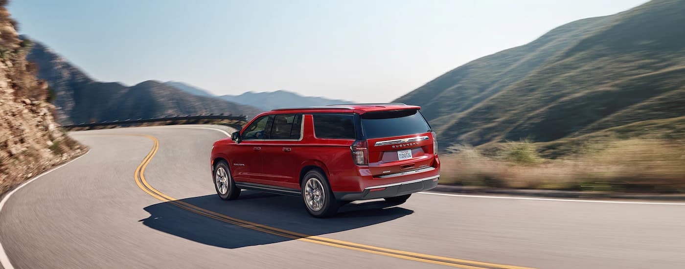 A red 2023 Chevy Suburban is shown from the rear driving on a mountain highway.