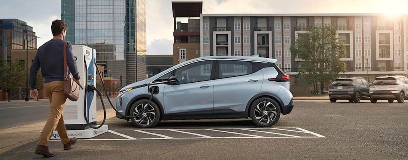 A man walks to a light blue 2022 Chevy Bolt EV plugged in at a public EV charging station.