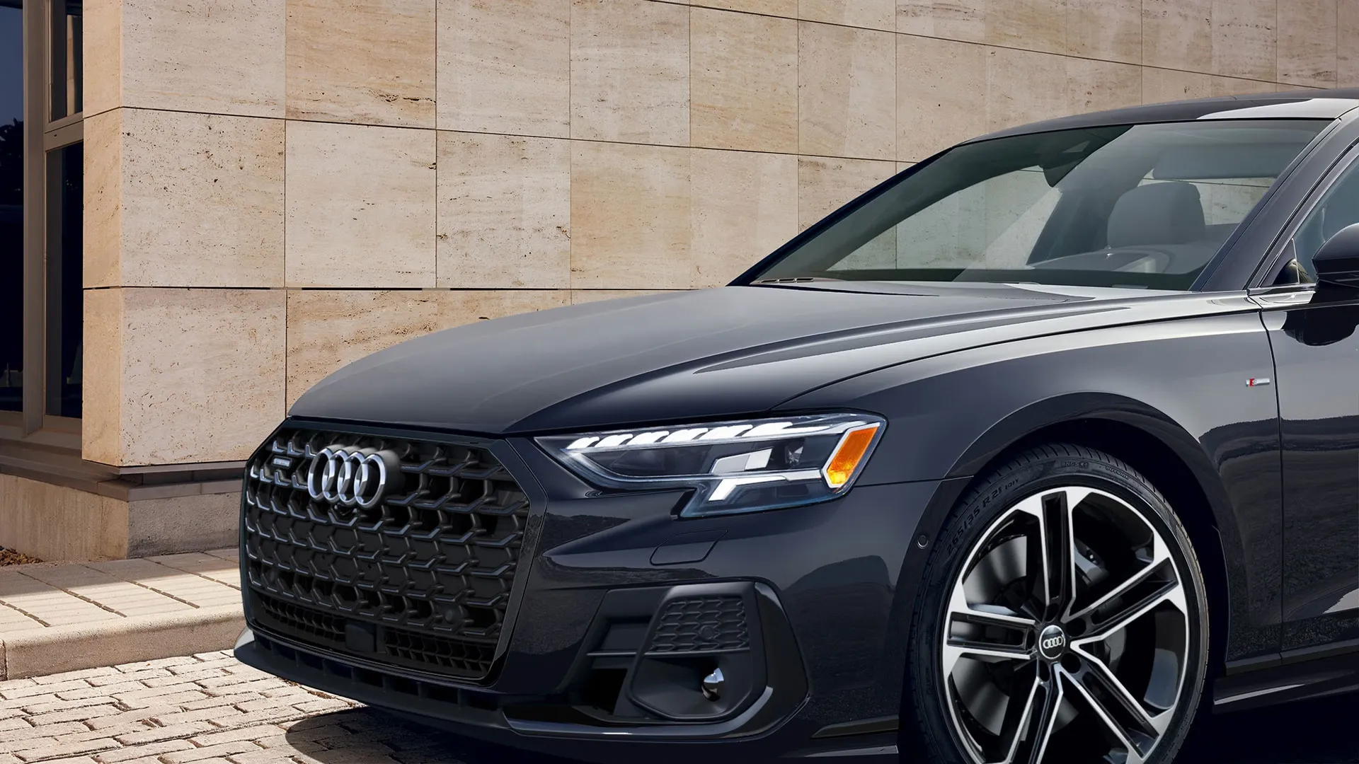 Vue rapprochée de la calandre noire et du phare gauche de l’Audi A8 2025 gris foncé stationnée sur pavés, montrant les jantes alliage bicolores et le logo Audi.