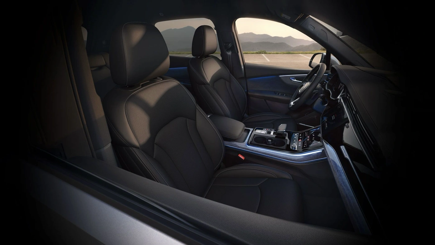 Habitacle de l’Audi Q7 2025 avec sièges avant en cuir noir, console centrale moderne et vue dégagée sur les montagnes au coucher du soleil.