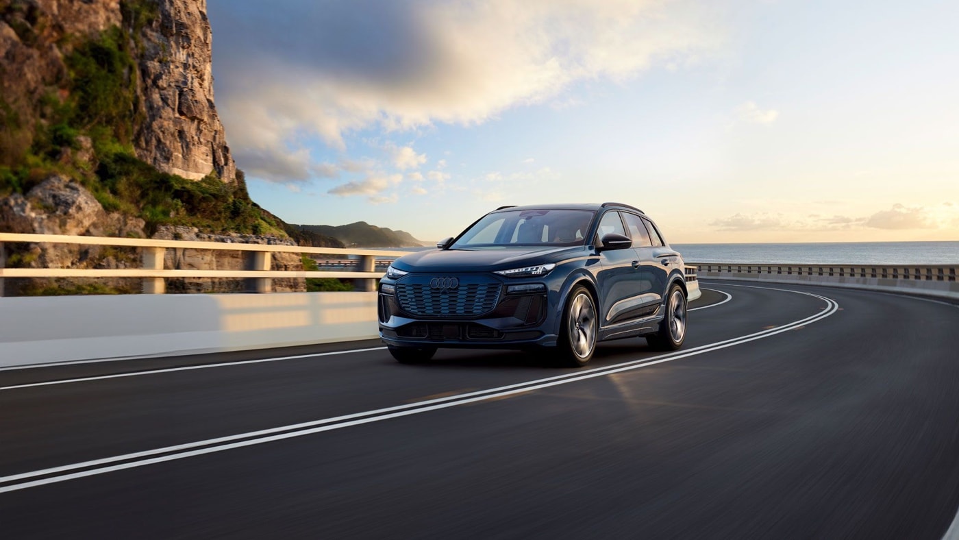 2025 Audi Q6 e-tron in dark blue driving along a coastal road at sunset, with a cloudy sky and cliffs in the background.