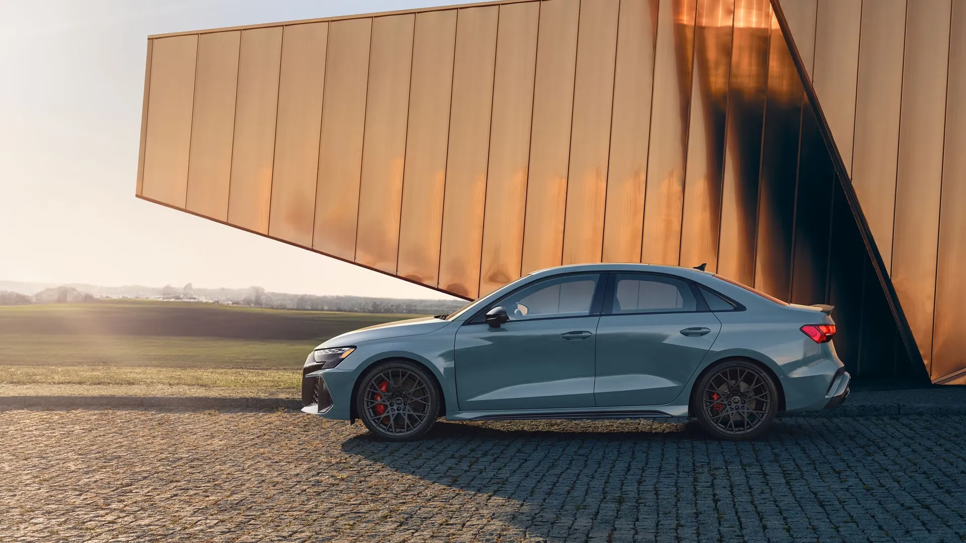 Audi RS 3 2025 bleutée vue de côté, stationnée sur un pavé en fin de journée devant un bâtiment doré.