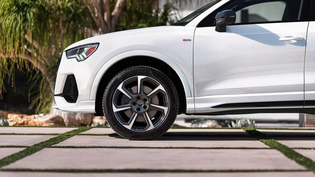 Close-up view of the front wheel and left fender of the white 2025 Audi Q3, parked on a modern driveway with tropical trees in the background.