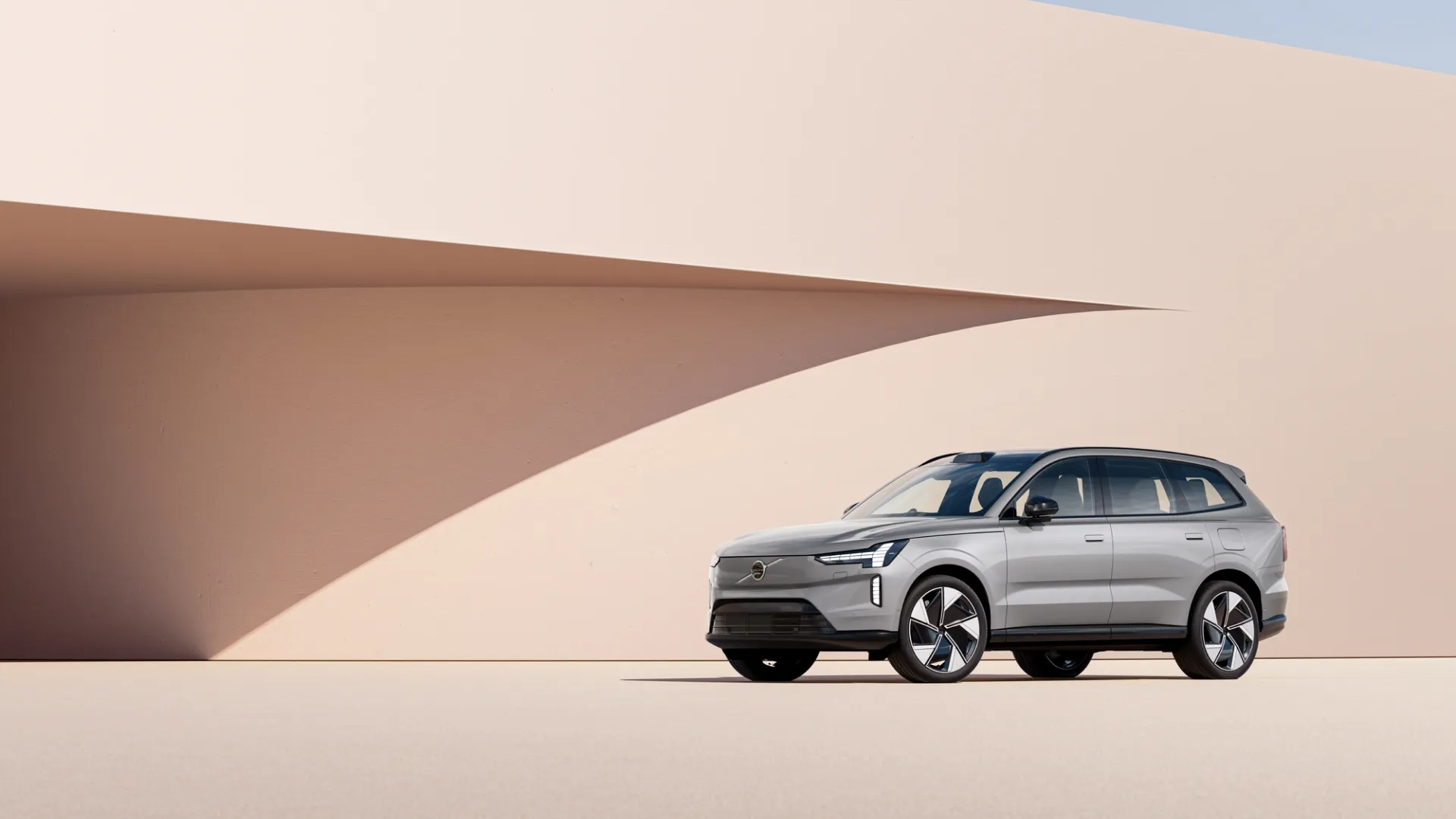 Volvo EX90 2025 silver parked in front of modern beige and pale pink architecture, illuminated by soft daylight.