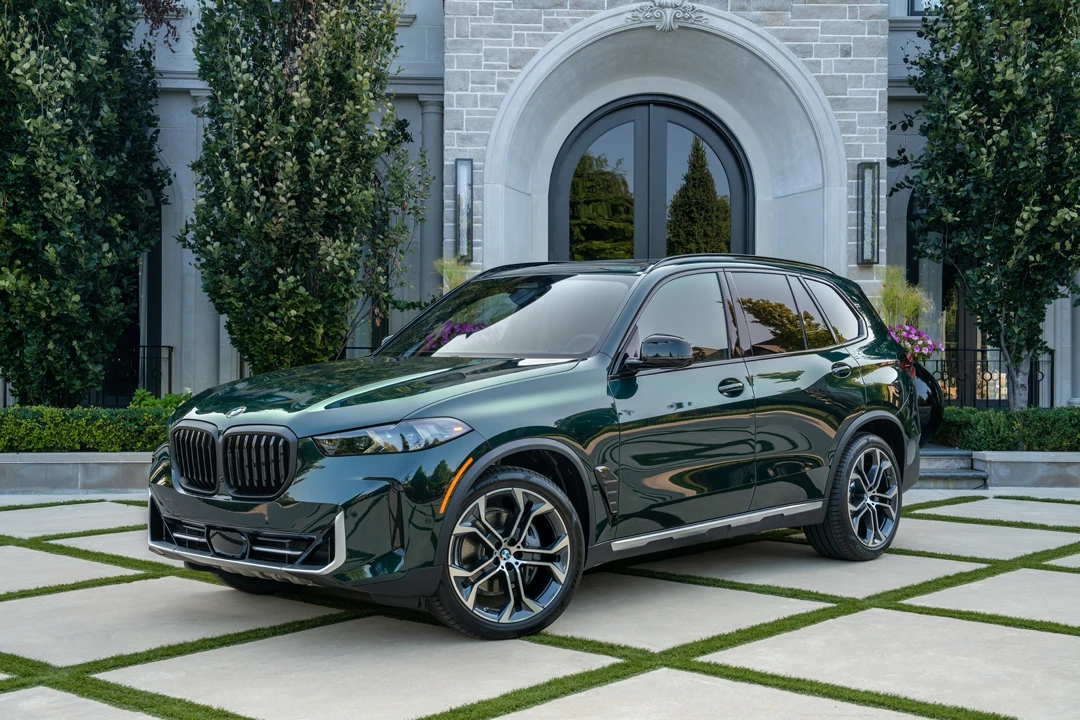 2025 BMW X5 dark green three-quarter front left view parked in front of a luxury house with landscaped garden.