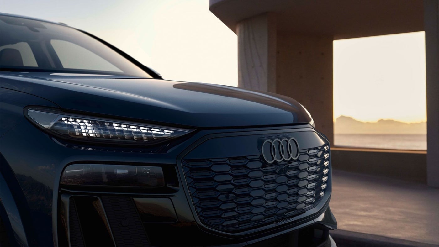 Close-up of the grille and left headlight of the 2025 dark blue Audi Q6 e-tron, Audi logo in the centre with sunset background.