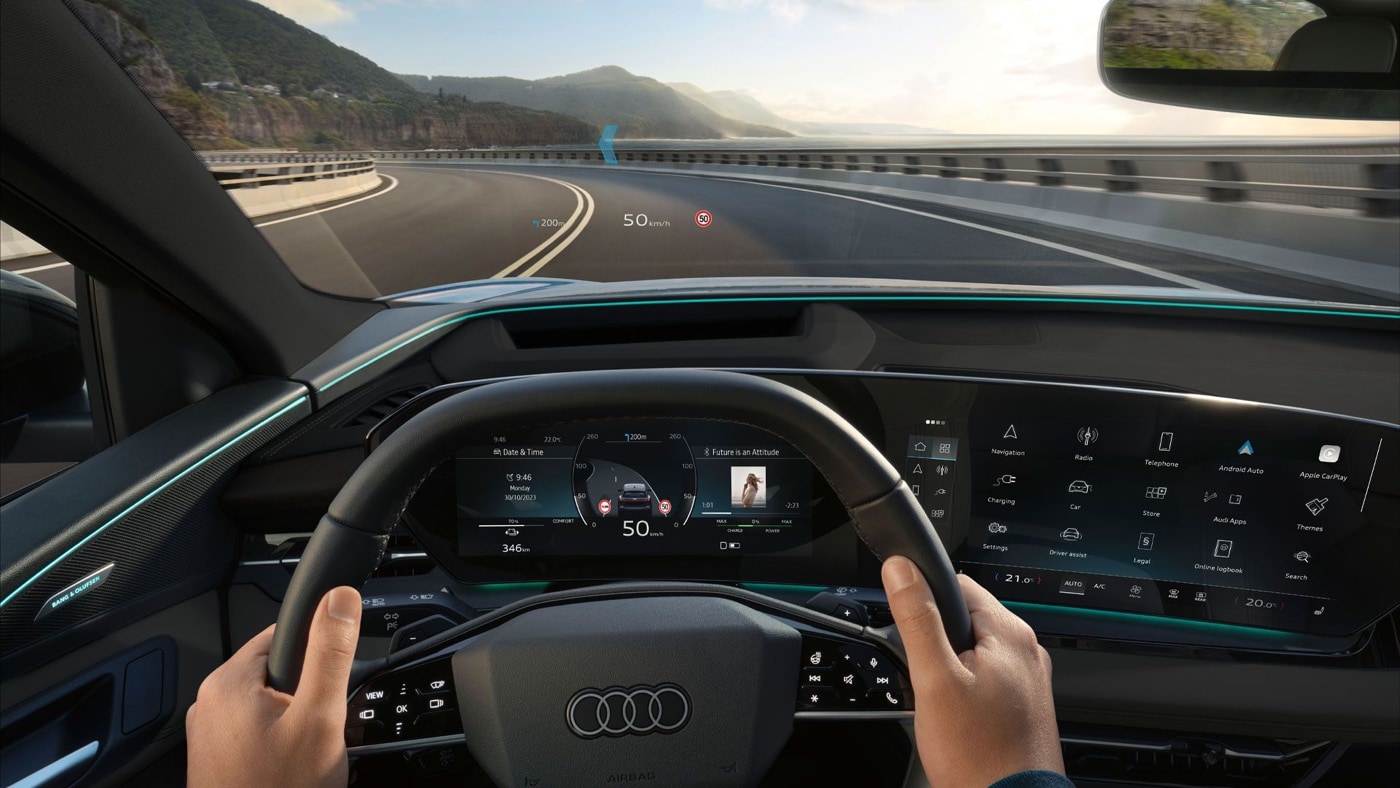Cockpit view of the 2025 Audi Q6 e-tron with Audi steering wheel, digital dashboard, and head-up display on panoramic road.