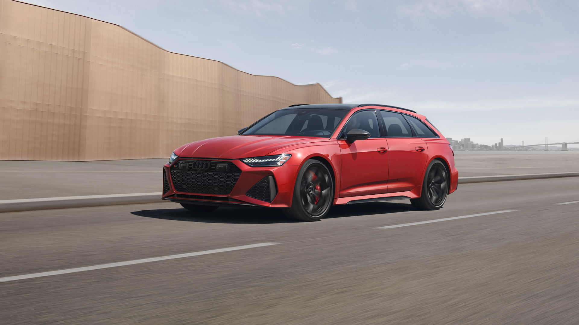 Red 2025 Audi RS 6 Avant driving on an urban road with an architectural wall in the background.