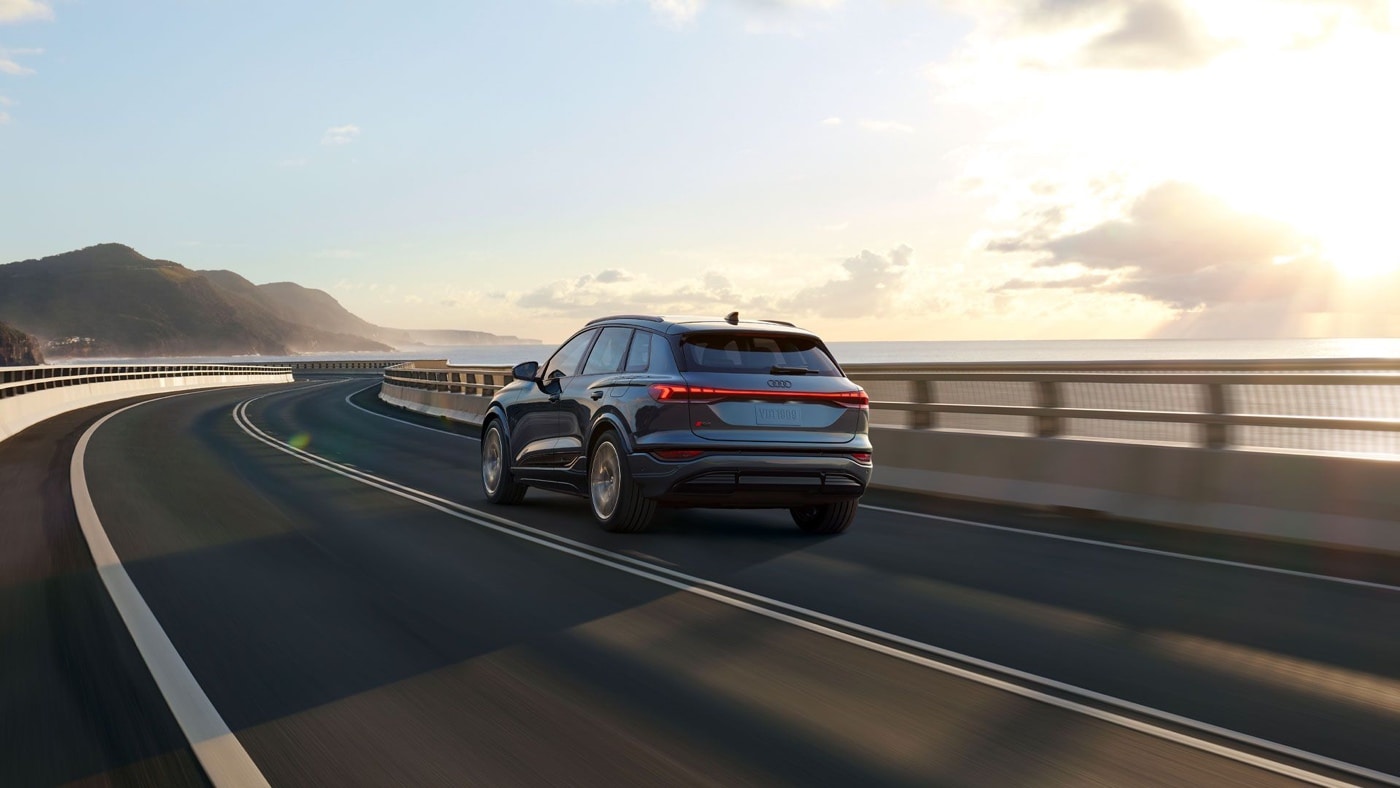 Rear view of the 2025 Audi Q6 e-tron driving on a coastal highway, electric SUV at sunset with ocean and mountains in the background.