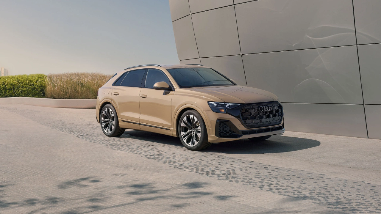 2025 Audi Q8 beige three-quarter front left view parked in front of modern metallic curved architecture.
