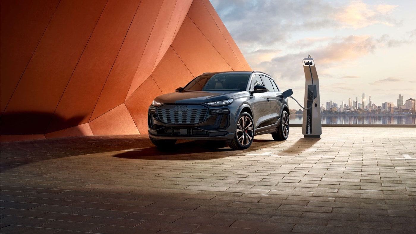 2025 black Audi SQ6 e-tron plugged into a fast-charging station, parked in front of an orange design wall with city skyline at sunset.