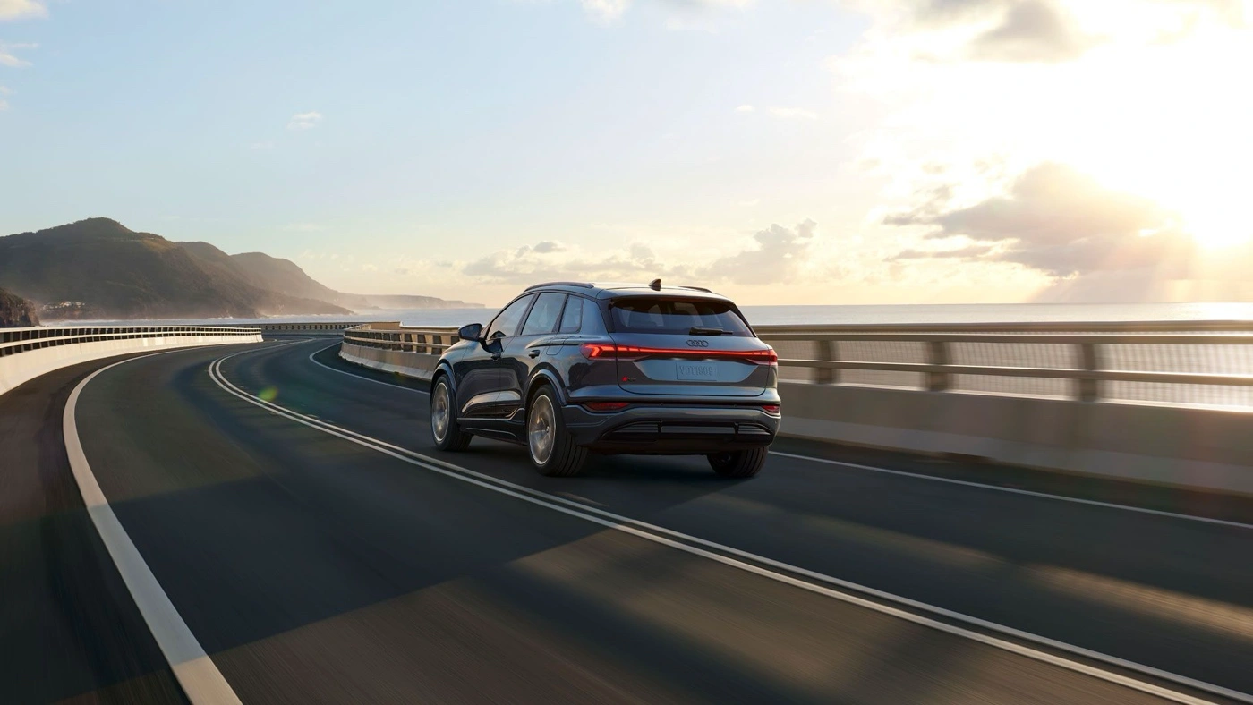 Audi Q6 e-tron 2025 bleu en trois quarts arrière sur pont côtier au soleil couchant, VUS électrique adapté au remorquage léger.