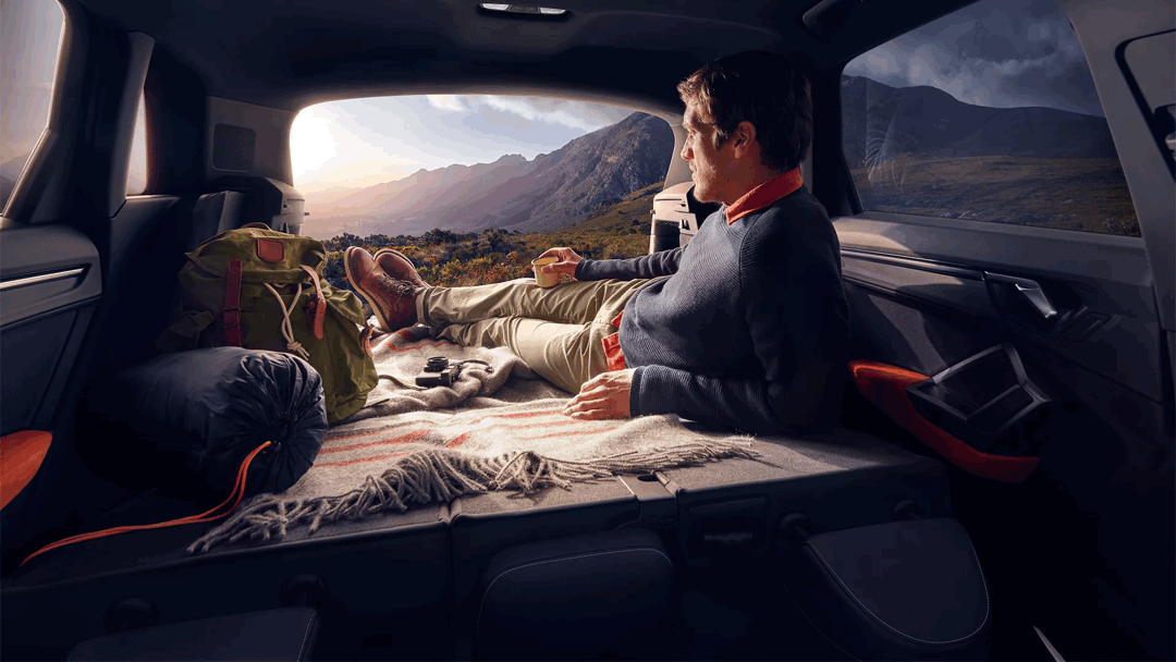 Person lying in the back of a 2025 Audi Q3 with the rear bench folded flat, looking out at a mountain sunset, with a hiking bag and blanket.