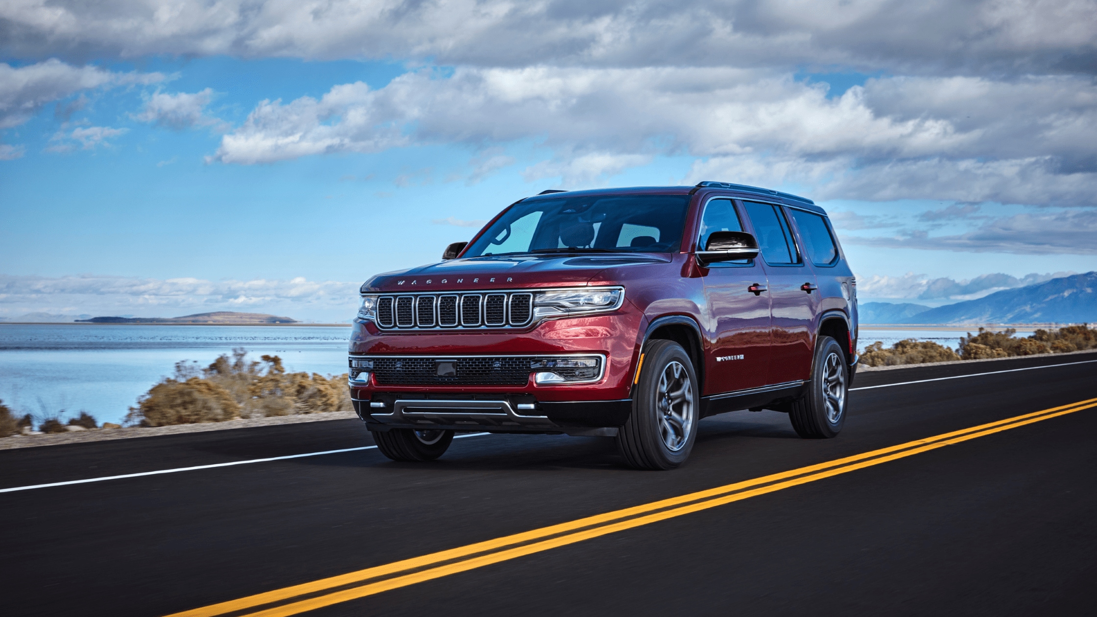 A red JeepÃÂ® Wagoneer driving on a scenic road with a lake and mountains in the background under a partly cloudy sky.