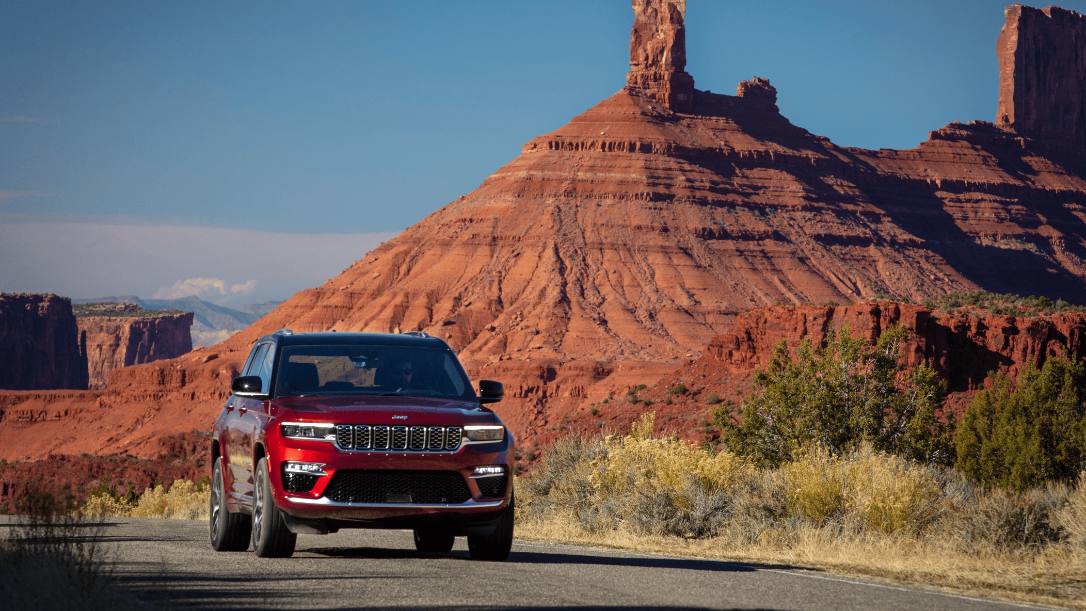 Red 2026 Jeep Grand Cherokee driving through a scenic desert landscape near Cape Coral, FL with towering red rock formations in the background.