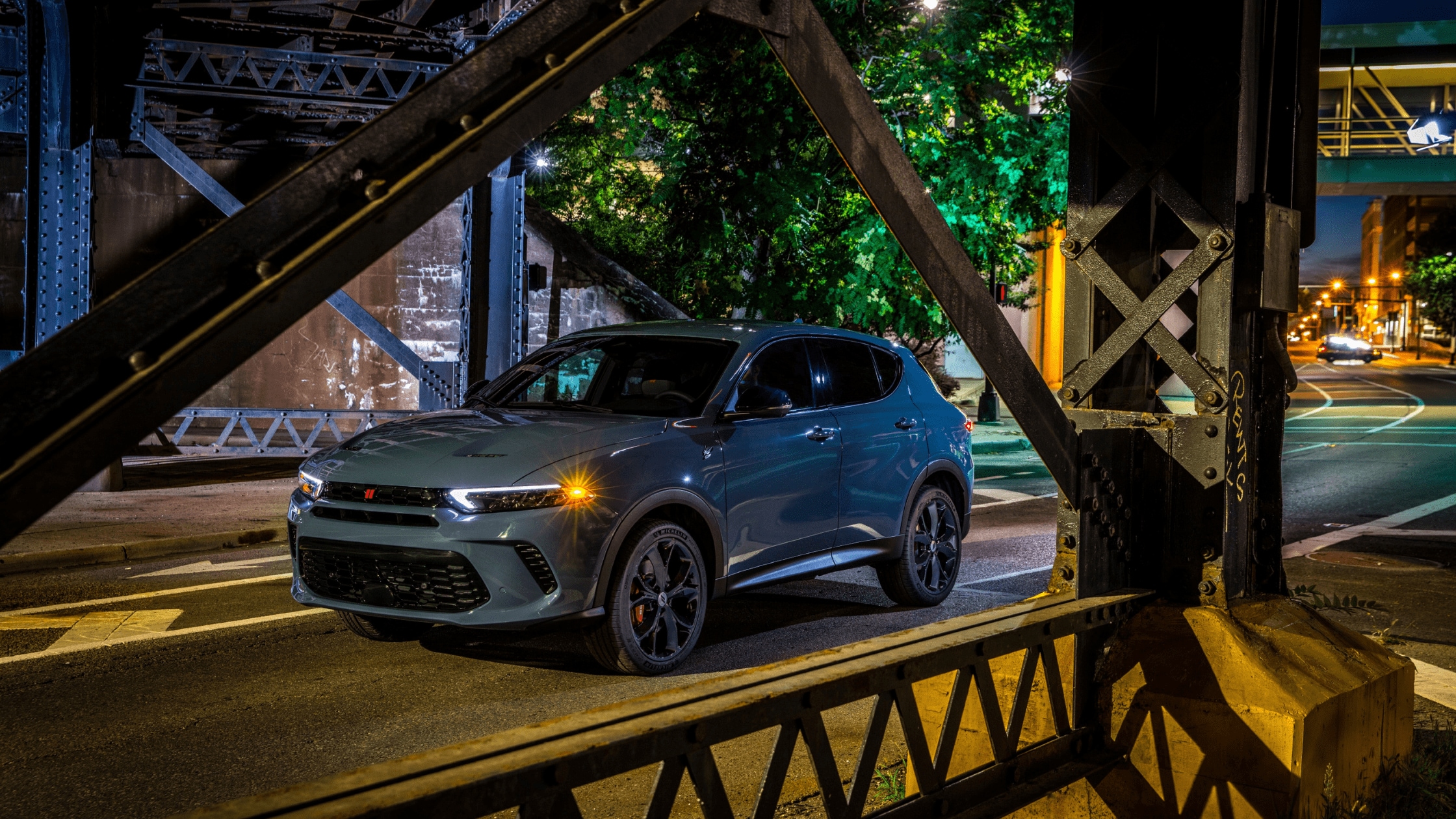 A 2024 Dodge Hornet is parked under a city bridge at night, illuminated by streetlights and surrounded by urban architecture.