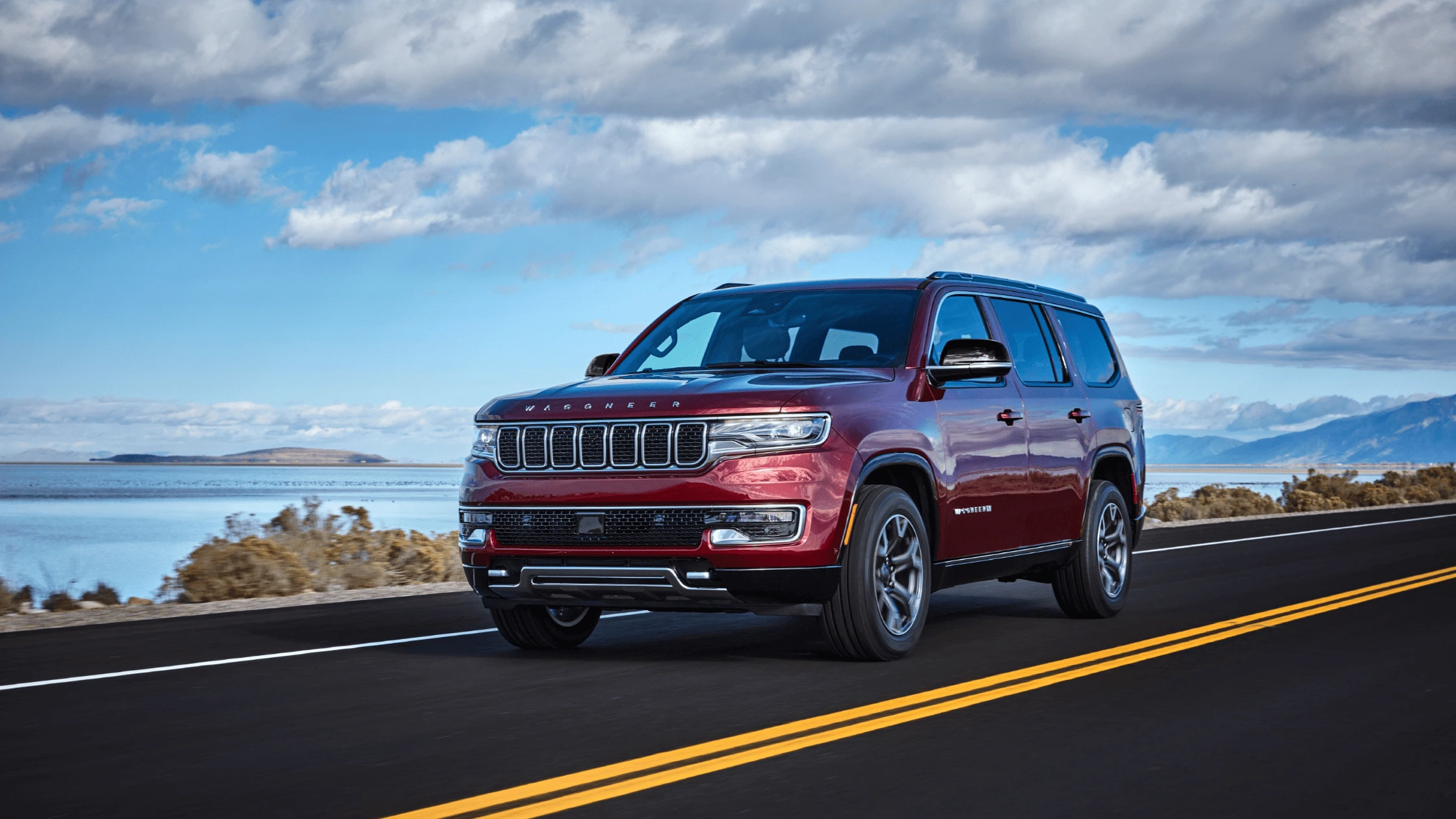 2024 JeepÂ® Wagoneer in deep red driving along a scenic lakeside road, showcasing its premium design and capability for outdoor adventures near Fort Myers.