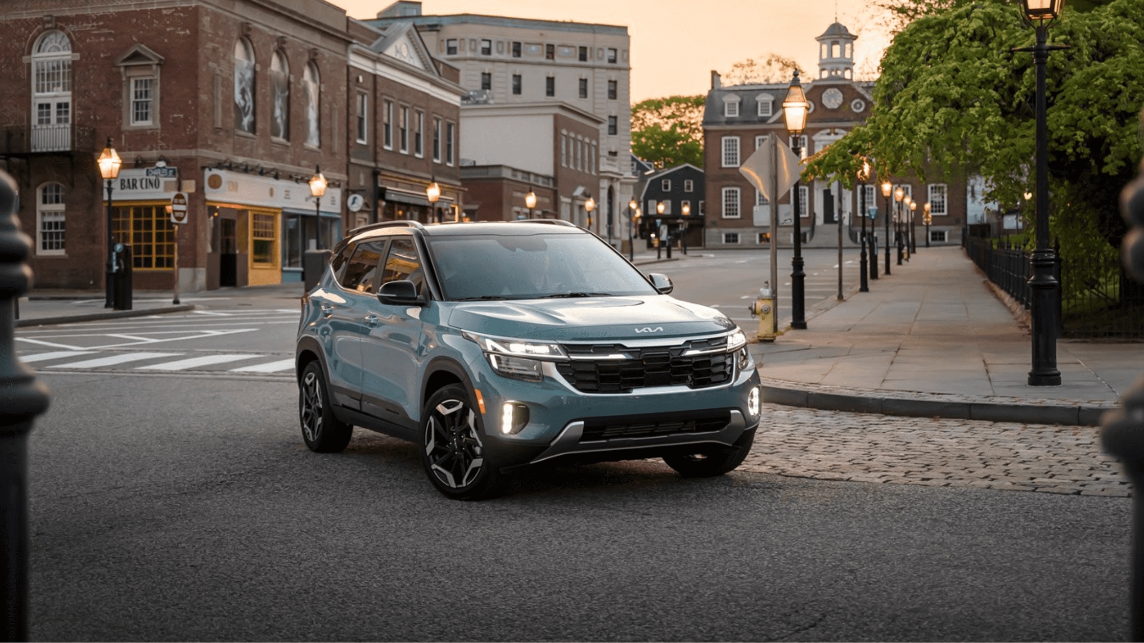 A light blue Kia Seltos parked on a charming cobblestone street in a historic town setting during the evening.