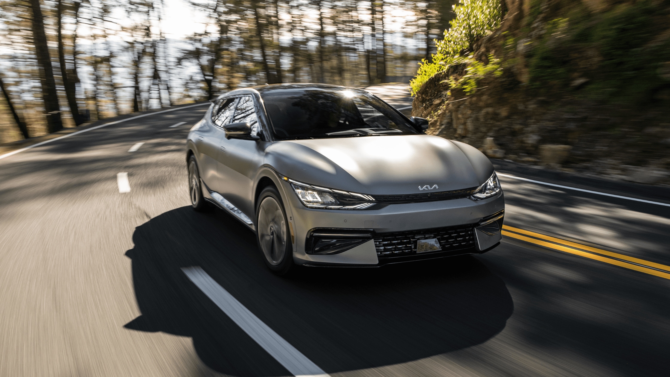 A silver 2024 Kia EV6 is driving on a winding forest road, surrounded by tall trees and dappled sunlight. The car's sleek design and modern headlights are prominently featured as it navigates the curves of the scenic route.