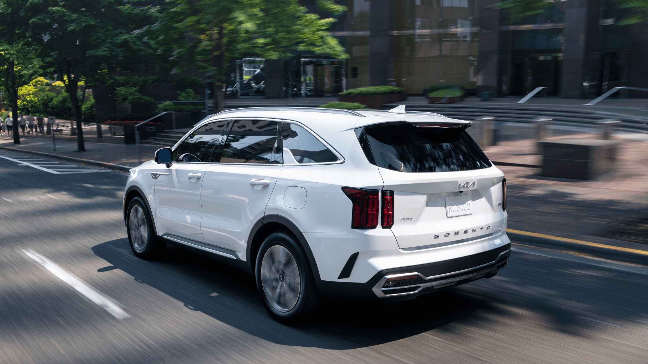 A white 2024 Kia Sorento driving on a city street, with blurred trees and buildings in the background, showcasing the vehicle's modern design and dynamic movement.