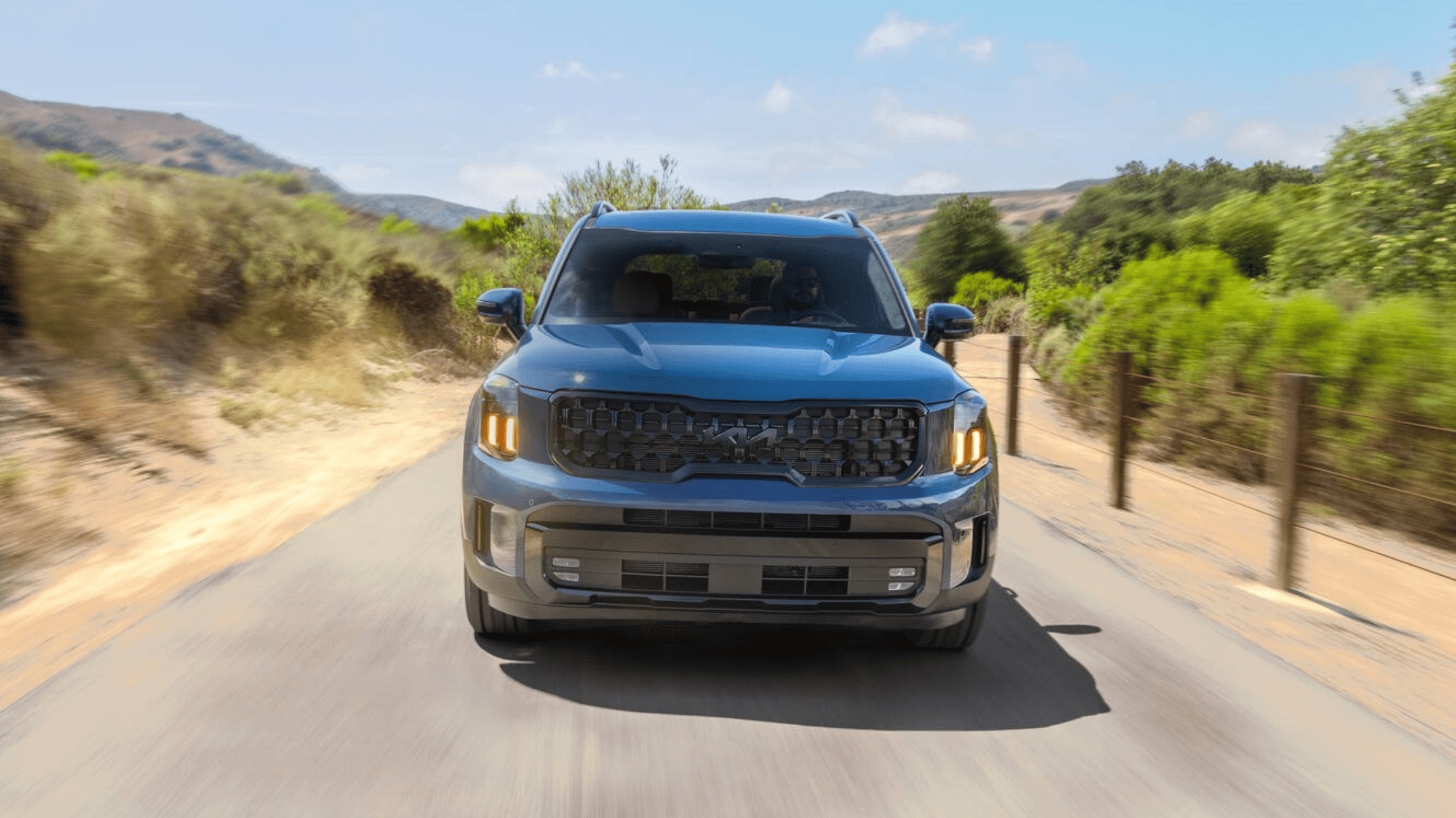 Front view of a blue 2025 Kia Telluride driving on a sunny scenic road near Cape Coral, FL with greenery and hills in the background.