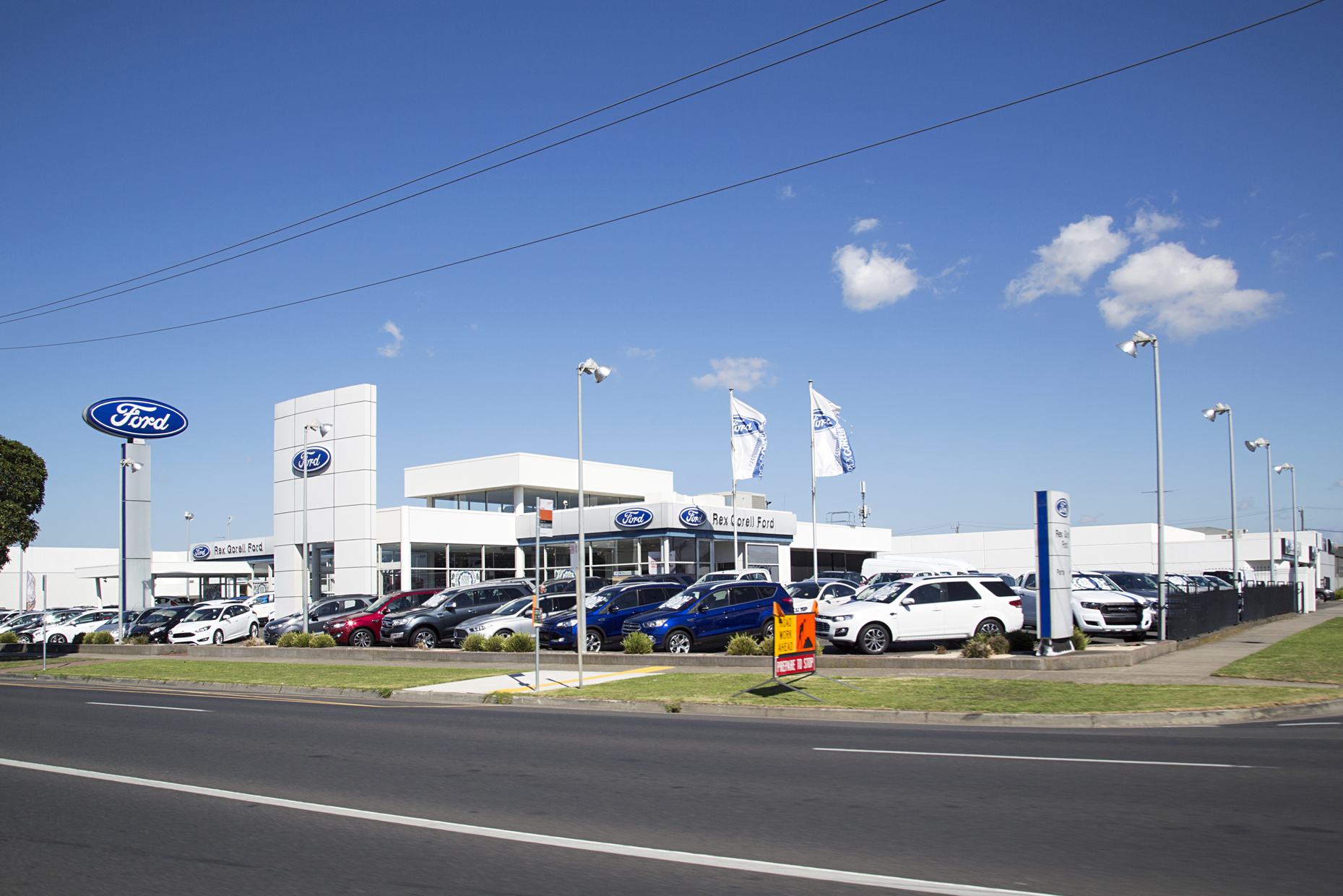Ford dealership showroom and vehicle inventory displayed along a roadside under a clear blue sky