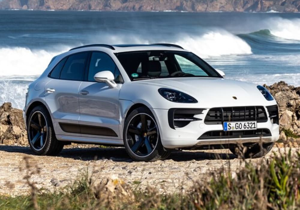 Front exterior passenger side of a white 2020 Porsche Macan parked by ocean water crashing over the rocky shore