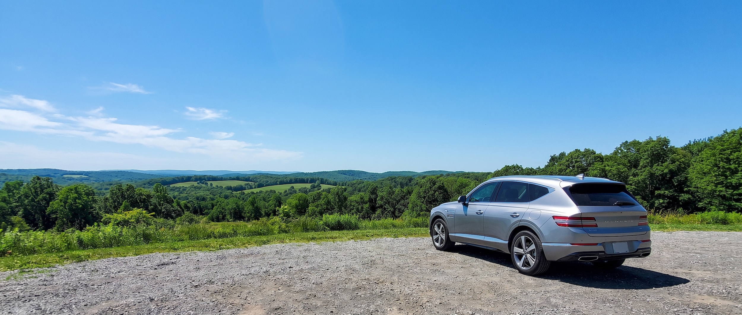 A 2026 Genesis GV80 in a light silver or white exterior parked at a scenic overlook with lush green Middle Tennessee rolling hills in the background under a bright blue summer sky, midday summer light.