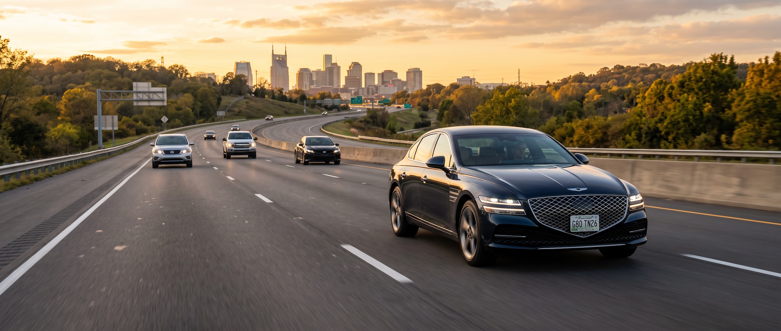 A 2026 Genesis G80 sedan in a refined dark exterior color on a Tennessee highway at golden hour, light traffic visible ahead, Nashville skyline faintly in the background, conveying a composed, confident commute