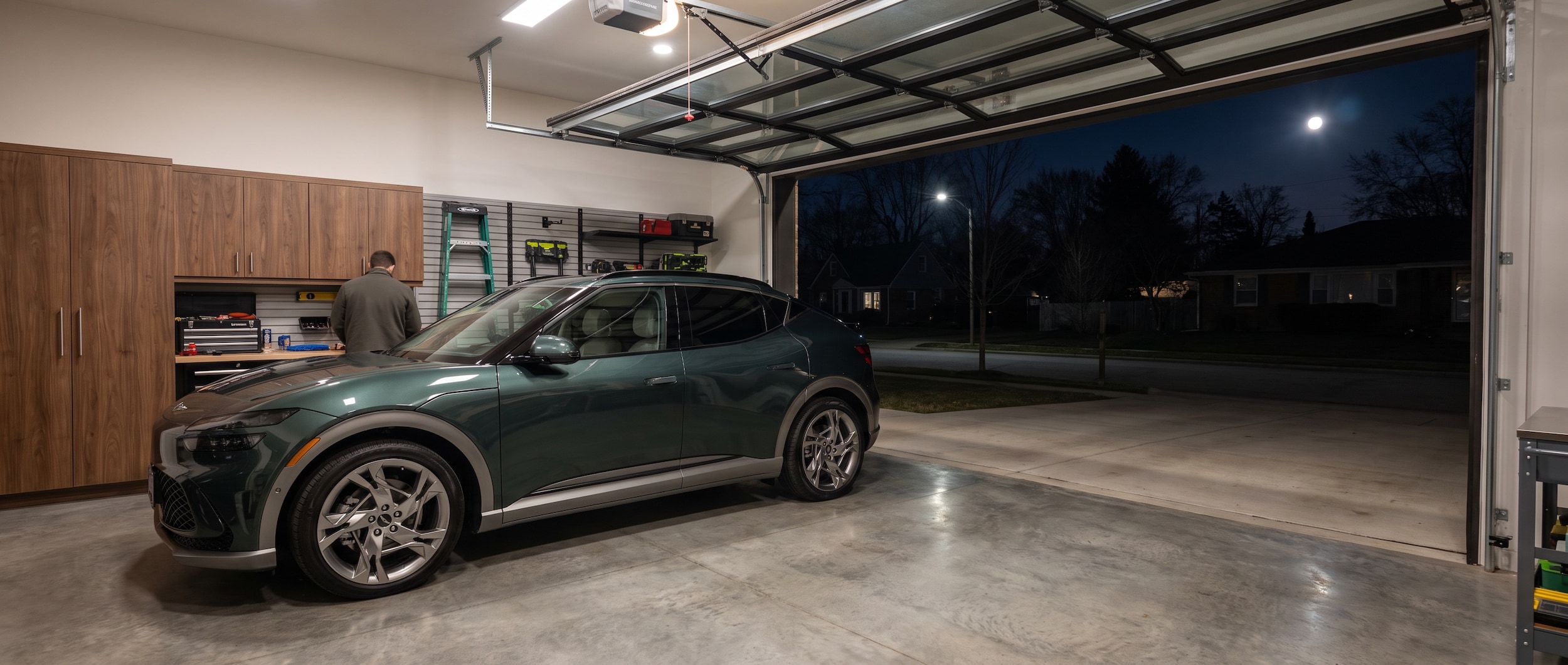 A 2026 Genesis GV60 in a modern suburban garage at night, plugged into a Level 2 wall charger with a soft blue charging indicator light glowing, garage door slightly open revealing a quiet Williamson County neighborhood
