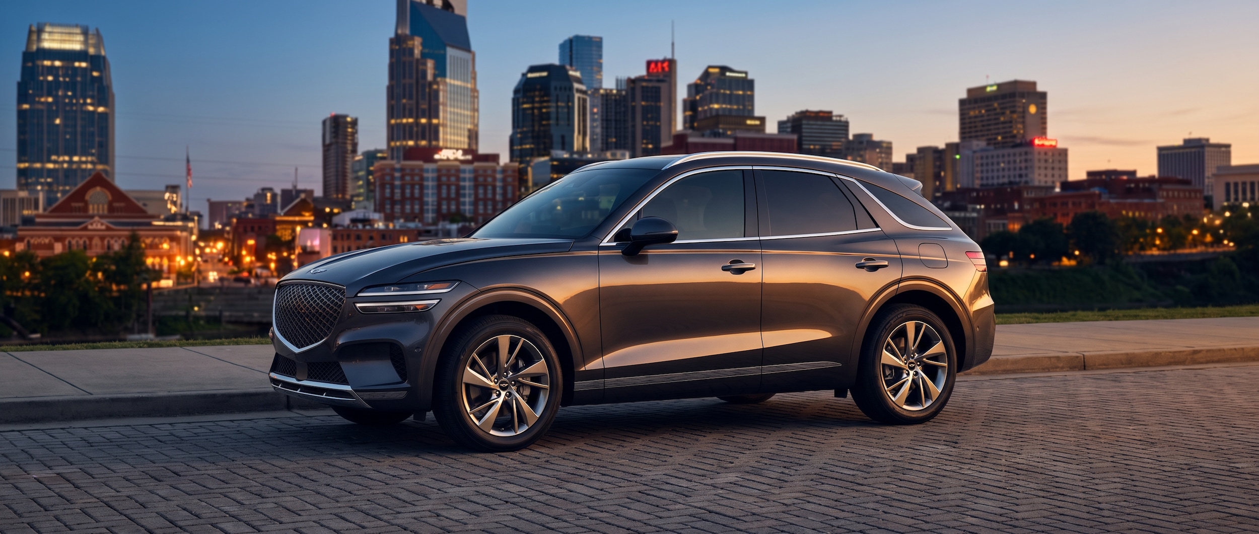 A 2026 Genesis GV70 in a deep navy or graphite finish parked on a clean urban street with Nashville's skyline visible in the background at dusk, studio-quality lighting catching the SUV's sculpted flanks and swept roofline, warm Tennessee summer evening atmosphere.