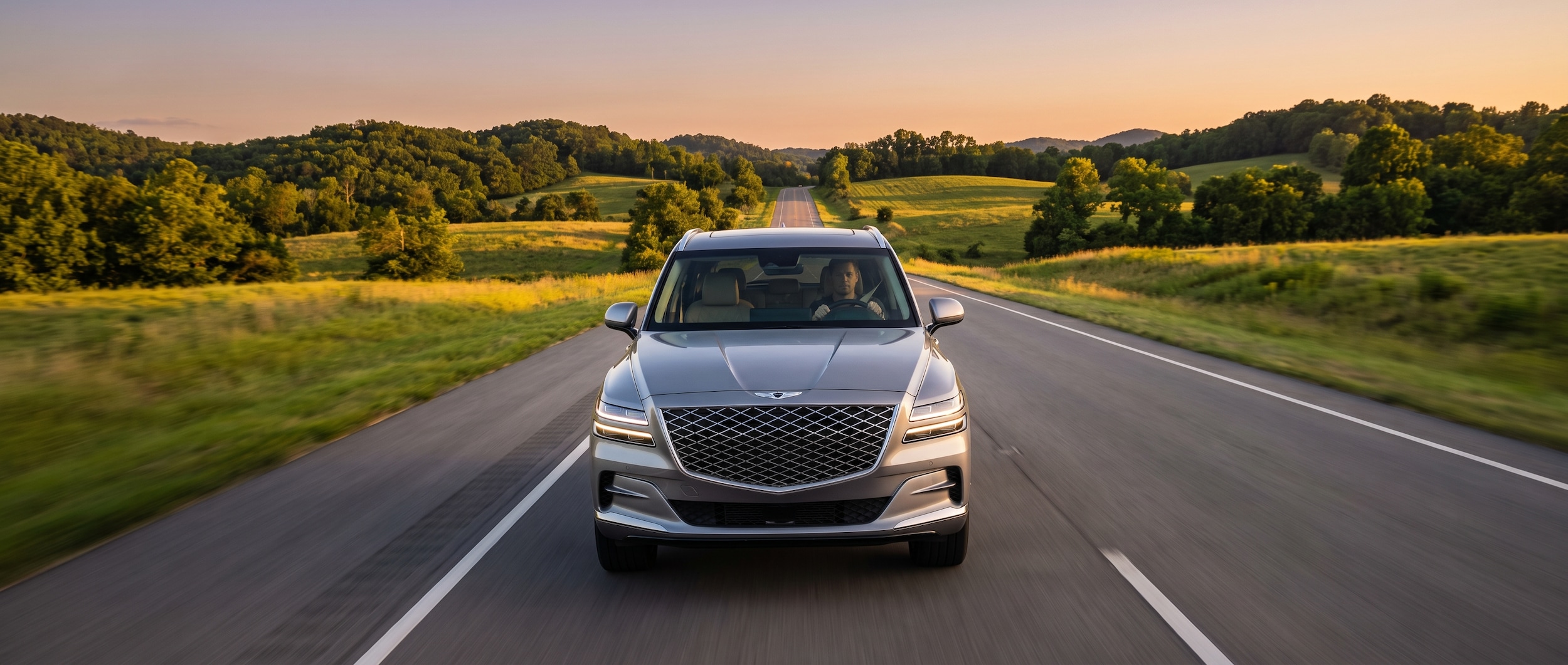 A silver Genesis GV80 on a wide Tennessee highway at golden hour, interior dashboard visible through the windshield, surrounded by rolling green hills and a clear open road stretching into the distance.