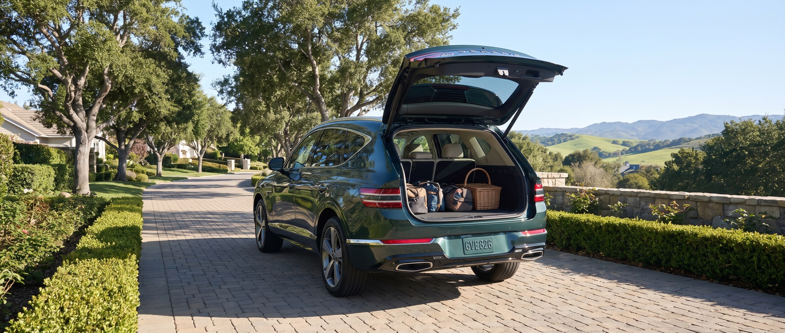 A 2026 Genesis GV80 in a sophisticated dark metallic finish parked in a tree-lined Brentwood, Tennessee residential driveway on a clear summer morning, rear liftgate open to reveal the spacious cargo area, green rolling hills visible in the background